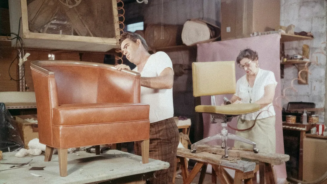 Interior de un taller de tapicería con una butaca retapizada en piel marrón y una silla de asiento amarillo en proceso de trabajo sobre caballetes.